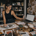 A woman in a black tank top and jeans sits at a wooden desk in a creative studio, sketching on paper and looking at a laptop screen surrounded by art supplies, books, and mixed-media materials.