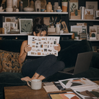 A woman sits on a dark velvet couch in a cozy, styled room, holding up a large printed sheet filled with small product photos. An open laptop rests on the couch beside her, with a mug on a wooden table in the foreground and shelves of books and decor in the background.
