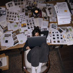A person sits in a chair at a cluttered wooden desk covered with papers, sketches, and design materials, holding a laptop from above.